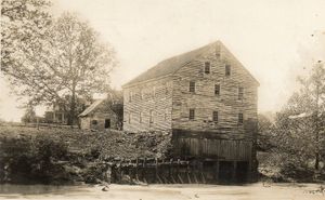 Black and white historic photo of large building along river with trees and small building in background.